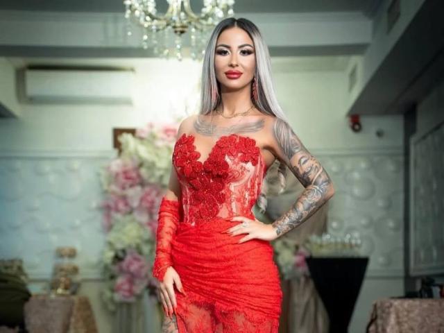 CelineDyor in a red lace dress, showing off her big boobs, tattooed arm, and long silver hair, standing with a hand on her hip, smiling.