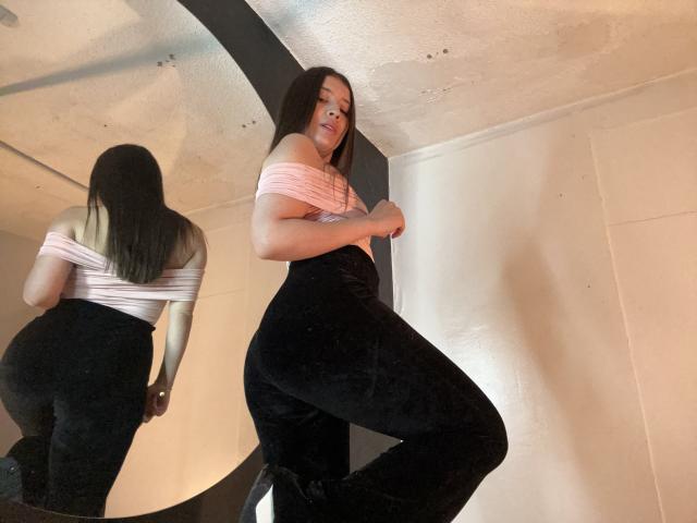 EmmaLombardi with long brown hair, pink off-shoulder top, and black velvet pants, standing near a mirror, showing her ass and reflection, in a slightly dirty room.