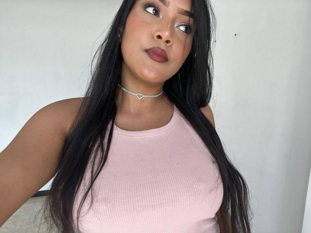 VickyRuiz mit langem, schwarzem Haar, die ein rosa Rippstrick-Tanktop, eine Herz-Kette und dunkelroten Lippenstift trägt, blickt zur Seite und hat leicht geschlossene Lippen. Brüste sind sichtbar..