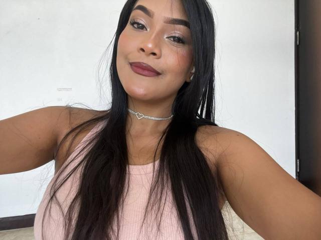 VickyRuiz mit langem, schwarzem Haar, einem pinken Tanktop und einer Diamantkette, lächelt in die Kamera. Sie hat braune Haut, dunkle Augenbrauen und pinken Lippenstift. Ihre Brüste sind mittelgroß..