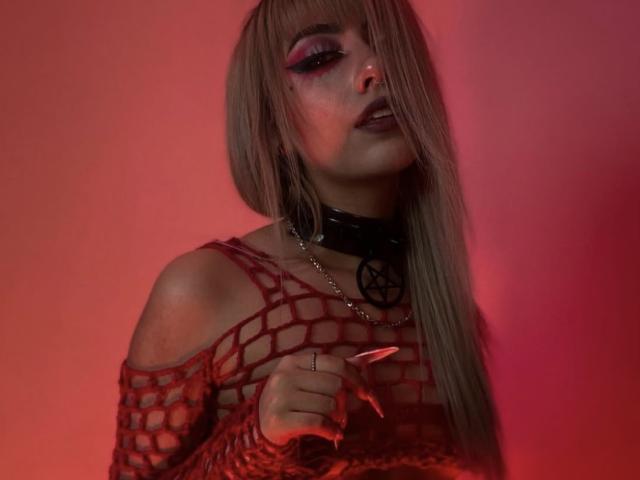 LitenKiitt a de longs cheveux blonds, un haut en filet rouge, un collier noir avec un pentagramme, du fard à paupières rouge, elle tient un couteau, l'éclairage est rouge, elle a une expression séduisante et regarde légèrement vers le bas..