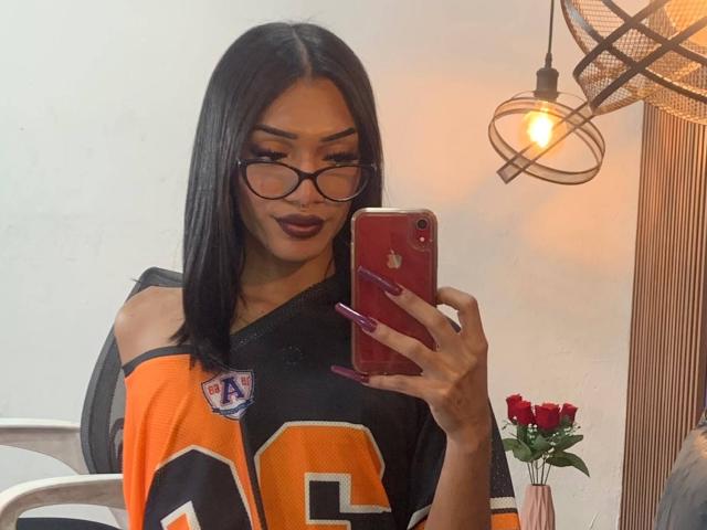 Debora Raquel, con cabello negro largo, gafas y lápiz labial oscuro, vestida con una camiseta naranja y negra de la NFL, tomando una selfie con un iPhone rojo, mostrando sus uñas moradas..