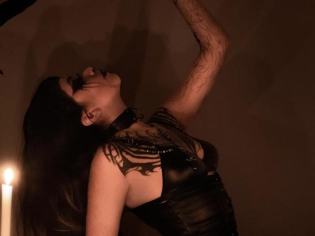 LitenKiitt con cabello oscuro, top de cuero negro y collar, brazos levantados, ojos cerrados, mirando hacia arriba, luz de vela en el fondo, tatuajes en el brazo, con sombras, con un ambiente melancólico, sensual..