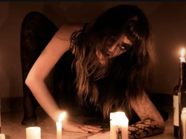 LitenKiitt, agachada, con cabello largo y castaño, medias de encaje negras y una blusa negra, alcanzando unas velas, con los brazos tatuados, en una habitación tenuemente iluminada, a la luz de las velas..