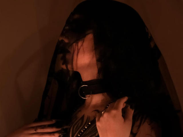 LitenKiitt com um véu de renda preta, usando um colar de couro preto com um anel de metal, mão no peito, iluminação marrom escura, renda semi-transparente, sombra facial visível..