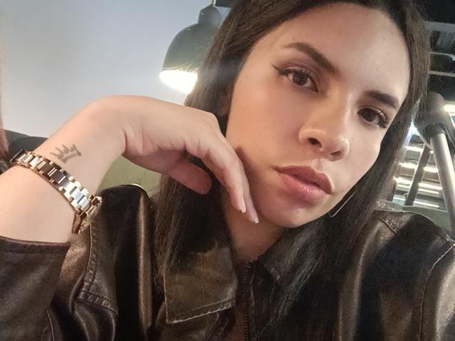 VioletaStone, com cabelo castanho escuro, vestindo uma jaqueta de couro, um relógio dourado e um anel, com o queixo apoiado na mão, mostrando uma pequena tatuagem no pulso esquerdo..