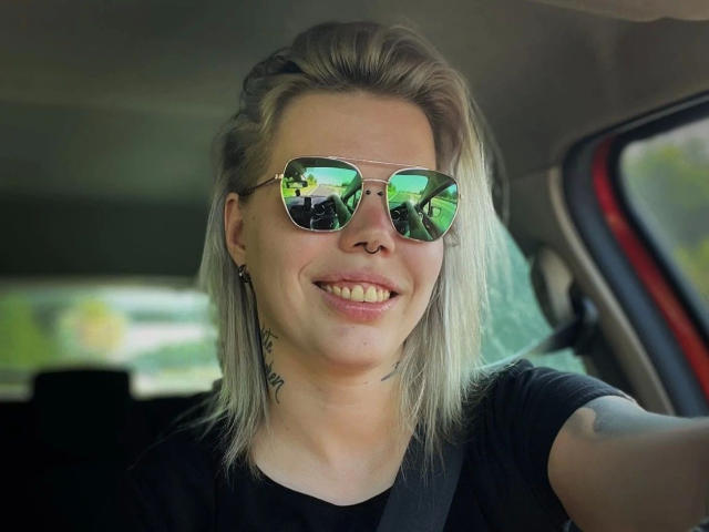 JuliaAvarix-hot with blonde hair, green reflective sunglasses, black t-shirt, smiling, visible nose ring, tattoo on neck, seatbelt, car interior, visible arm tattoo, relaxed, confident.