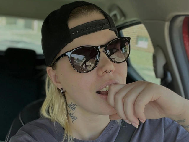 JuliaAvarix: atractiva, con cabello rubio, gorra negra puesta hacia atrás, gafas de sol negras, piercing en la nariz, tatuajes, camisa gris, dedo en la boca, interior de un coche, sexy, atractiva, informal..