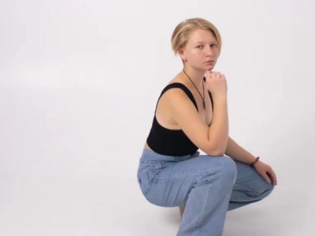 Laila Lafreniere sitzt im Hocke, trägt ein schwarzes Tanktop und blaue Jeans, hat kurze blonde Haare und einen ernsten Gesichtsausdruck, ihre Hand ist in der Nähe ihres Kinns..