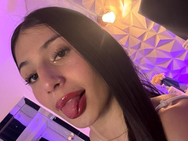SophieBeaumont's face with long black hair, shiny wet lips, nose ring, and tongue out, in a purple-lit room, showing her glossy eyes and light skin.