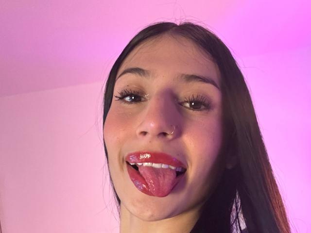 SophieBeaumont has a nose ring, long black hair, and is sticking out her tongue with shiny red lips, her tongue has a white sticker. Pinkish light highlights her face.