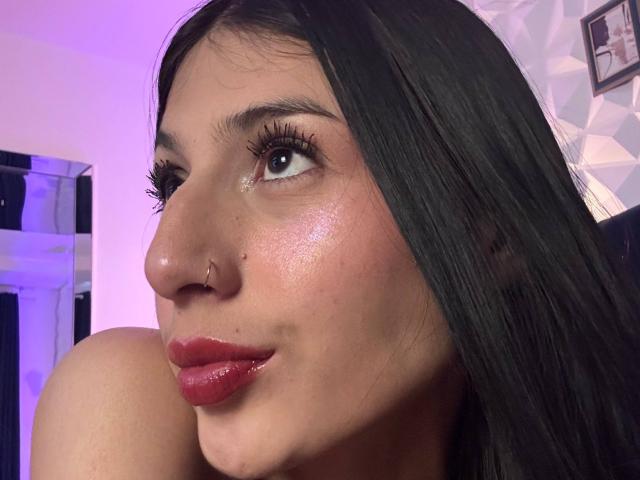 SophieBeaumont's face with glossy red lips, shiny skin, black eyelashes, nose piercing, and black hair. She's looking to the side with a slight smile. Boob partially visible.