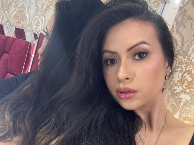 HannaRobertss has shiny pink lipstick, long wavy black hair, and is staring at the camera with dark, slightly arched eyebrows, wearing a black top. Her face is centered.