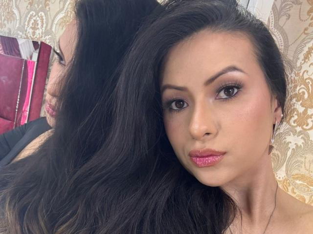 HannaRobertss, avec ses longs cheveux noirs, son rouge à lèvres pailleté rose et ses yeux marron, a l'air sérieuse sur une photo d'elle-même, montrant son visage et sa partie supérieure de la poitrine, et porte un collier..