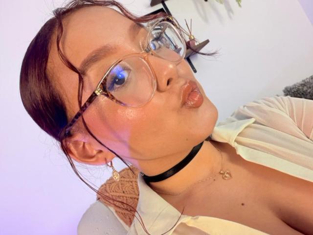 Sophia Browm, con labios carnosos, usando gafas, un collar negro y una camisa beige, tiene el pelo castaño ondulado y piel clara, mostrando un escote sutil, y lleva un collar en la foto..