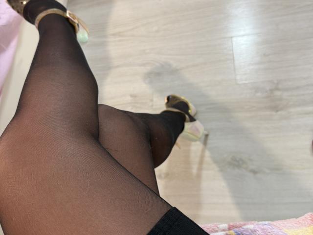 De zwarte, doorschijnende panty's van RoxanneQuin, gekruist bij de enkels, laten haar zwarte stilettohakken met gouden enkelbandjes zien op een houten vloer. Haar voeten wijzen omhoog..