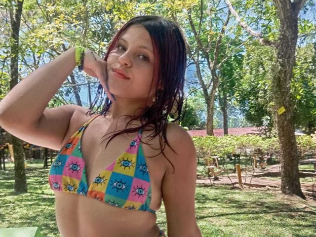 SophieHill with wet purple hair, wearing a colorful checkered bikini top, has small tits, and a green wristband, standing in a sunny park.