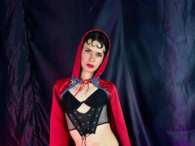 JazmiinRosee met een rode capuchon, een zwarte beha, netkousen en een rode cape, die kleine borsten en een schattig gezicht laat zien, en die staat voor een donkere achtergrond. Ze draagt rode lippenstift..