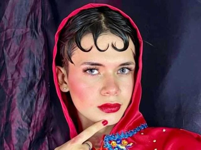 JazmiinRosee, com cabelo preto curto, capuz vermelho, batom vermelho, olhos azuis e unhas vermelhas, apontando para o queixo, vestindo um vestido vermelho com bordados azuis, olhando para o lado..