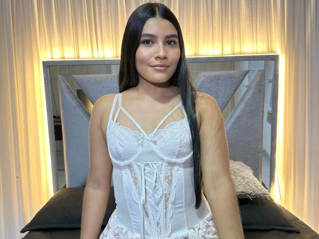 Catagomez with long black hair, wearing a white lace-up corset, has small tits, cute face, and a subtle smile, sitting on a bed with black pillows.