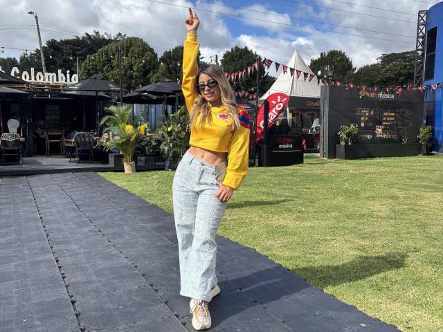 AmaraRicci, with long blonde hair, wearing a yellow crop top, and light blue high-waisted jeans, raises one finger, standing on a blue tiled path, in a sunny yard.