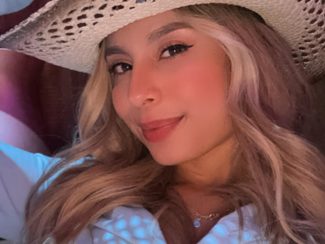 AmaraRicci has blonde, wavy hair, wearing a white hat and a blue top. She's smiling with red lipstick, black eyeliner, and a small necklace. Her face is slightly flushed.