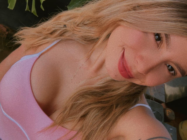 AmaraRicci has blonde wavy hair, pink tank top, and is smiling with red lipstick. Her medium-sized tits are visible, showing a cute and sexy look.