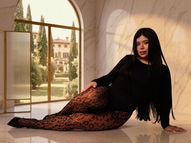 Chloe, com cabelos longos e pretos, blusa preta de manga comprida e collant com estampa de cobra, apoiada em um piso de mármore, usando sapatos pretos, olhando para a câmera..