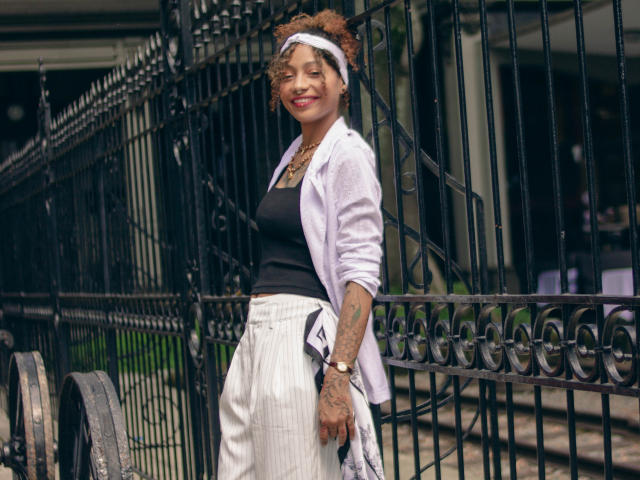 DaianaCurly with curly brown hair, white headband, black tank top, white pants, pink cardigan, tattoos on right arm, smiling, standing by black iron fence, holding patterned cloth.