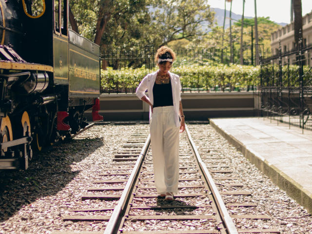 DaianaCurly, met krullend haar, een zwarte top, witte broek en een zonnebril, staat op treinrails, met een wandelstok in haar hand, naast een zwarte trein op een zonnige dag..