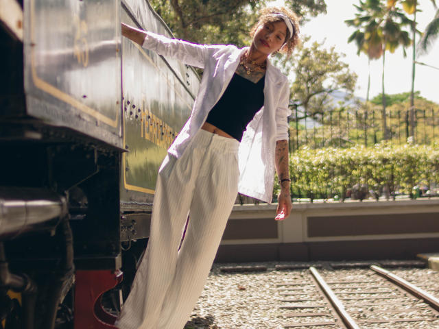 DaianaCurly est appuyée contre un train, son dos légèrement incliné, vêtue d'une chemise blanche, d'un haut noir et d'un pantalon blanc à rayures fines, laissant apparaître des tatouages et un collier. La lumière du soleil met en valeur sa silhouette..