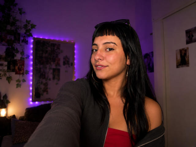 Kaliophe, con i capelli neri, indossa una giacca grigia e una maglietta rossa, sorride alla fotocamera, allungando un braccio, con luci viola sullo sfondo, un piccolo piercing al naso, orecchini a cerchio..