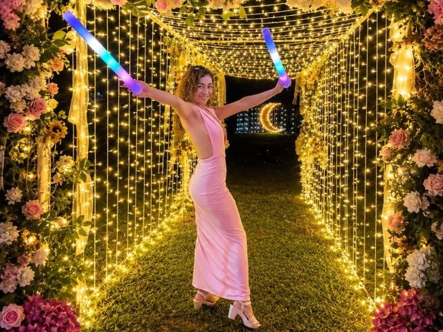 Arianna Blake trägt ein pinkes, ärmelloses Kleid und hält leuchtende blaue und violette Lichter in den Händen, die Arme sind ausgestreckt. Sie steht in einem Tunnel aus Blumen und Lichterketten, lächelt und hat lockiges Haar. Sie trägt High Heels..