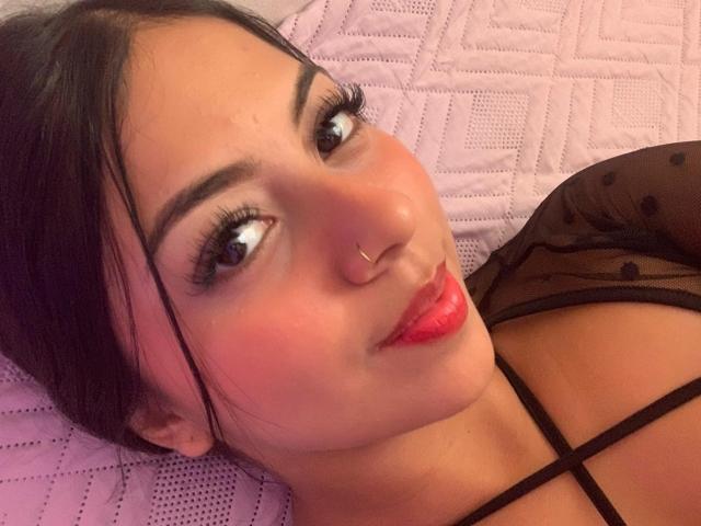 JuliethVelvett, con i capelli neri, il rossetto rosso, il piercing al naso e la maglietta nera a pois, sdraiata su un letto rosa trapuntato, mostra il seno e il suo viso con le labbra piene e le lunghe ciglia..
