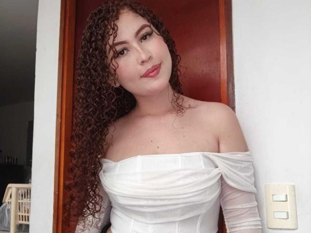 IrisDalton with curly brown hair, white off-shoulder dress, and pink lipstick, standing in front of a red door, smiling at the camera, showing her tits.