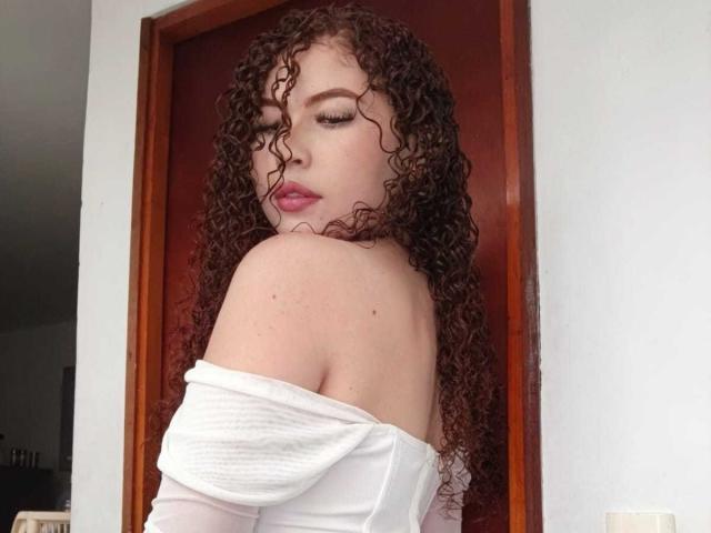 IrisDalton with curly red hair, white off-shoulder top, pink lipstick, and eyeliner, looking back over shoulder, showing bare shoulder and upper back.