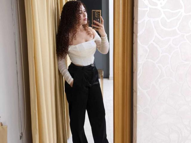 IrisDalton with curly red hair, white off-shoulder top, black high-waist pants, holding phone in mirror, hand in pocket, large boobs, confident stance.