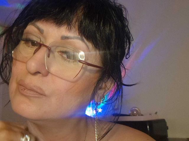 YummyTitss with black messy hair, glasses, and brown lips, wearing a star-shaped blue earring, looking slightly to the side, with colorful light reflections on her face.