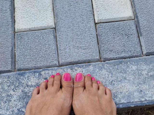 YummyTitss' Füße mit rosa lackierten Zehennägeln auf einem grauen, strukturierten Steinpflaster. Ihre Zehen sind leicht gespreizt, wobei ihre rosa lackierten großen Zehen sichtbar sind..