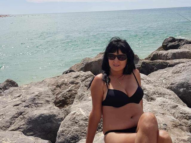 Leckere Brüste mit schwarzem BH und Unterhose, sitzend an einem felsigen Ufer, mit Sonnenbrille, lächelnd, mittelgroße Brüste, schwarzes Haar, gebräunte Haut, goldene Halskette und schwarzer Bikini-Unterteil, Ozean im Hintergrund..
