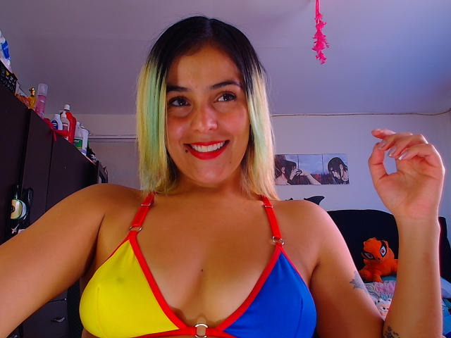 LaSexyColombiana, avec des cheveux dégradés blonds à verts, souriante, portant un soutien-gorge jaune, bleu et rouge avec des bretelles rouges, montrant sa poitrine généreuse, tenant ses cheveux, dans une chambre..