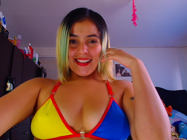 LaSexyColombiana met haar dat overloopt van blond naar groen, een rode, gele en blauwe top met bandjes, lachend, met een gemiddelde borst, rode lippenstift en een kleine tatoeage op haar rechterarm..