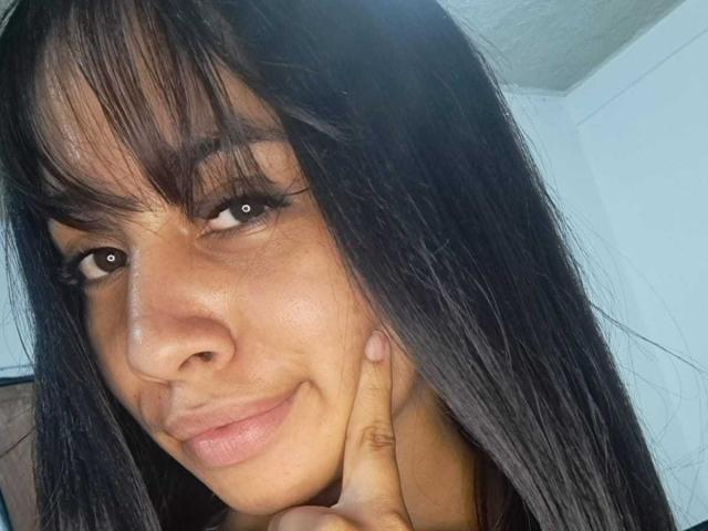 LovelyColombiaAngel com cabelo castanho escuro e liso, franja, olhos castanhos e um leve sorriso, tocando o queixo com o dedo direito, mostrando seu nariz delicado e lábios cheios..