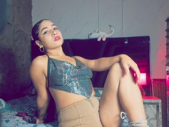 Una bellissima donna colombiana, con la pelle abbronzata, rossetto rosso e capelli scuri raccolti in uno chignon, indossa una top blu scintillante ricoperta di paillettes e pantaloncini beige, seduta su un letto con le gambe incrociate..