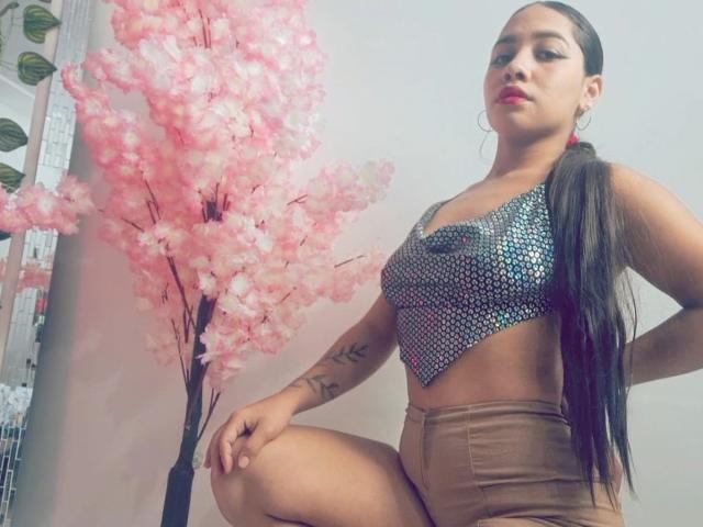 Una bellissima ragazza colombiana, con una lunga coda di capelli castani, indossa un top corto scintillante e pantaloncini beige, con le ginocchia scoperte, mostrando piccoli seni e un braccio tatuato, in piedi accanto a un albero di ciliegio rosa..