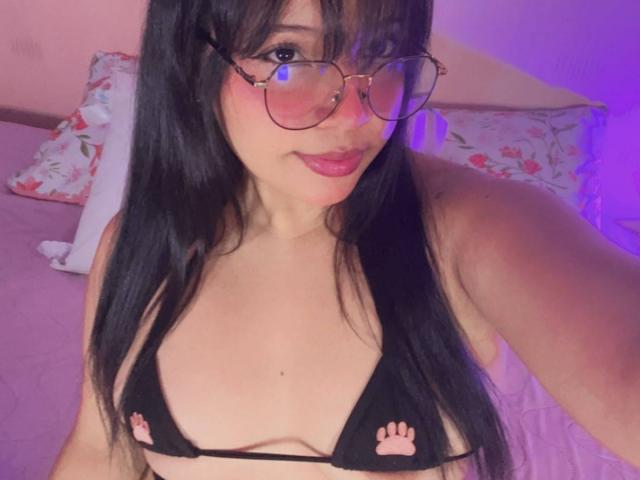 InnocenthaZel heeft zwart haar, draagt een bril en heeft een zwart bikini-topje aan met roze pootafdrukken, waardoor haar kleine borsten zichtbaar zijn. Ze zit op een roze bed met bloemenkussens..