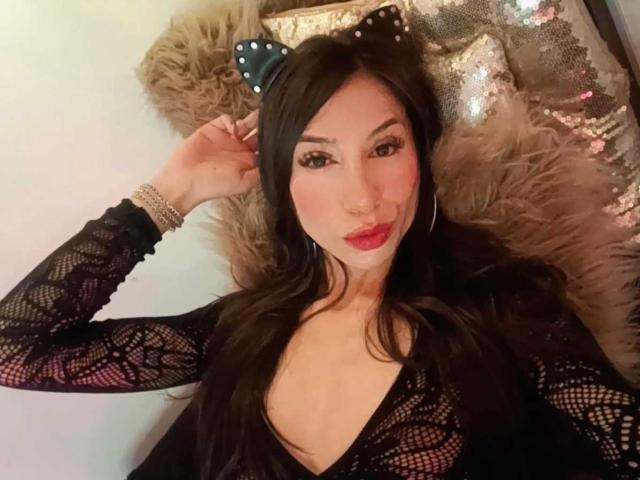 NahomyCarther with black lace top, red lipstick, and cat ears, lying on fur, showing her tits, wearing a bracelet, and looking at the camera.