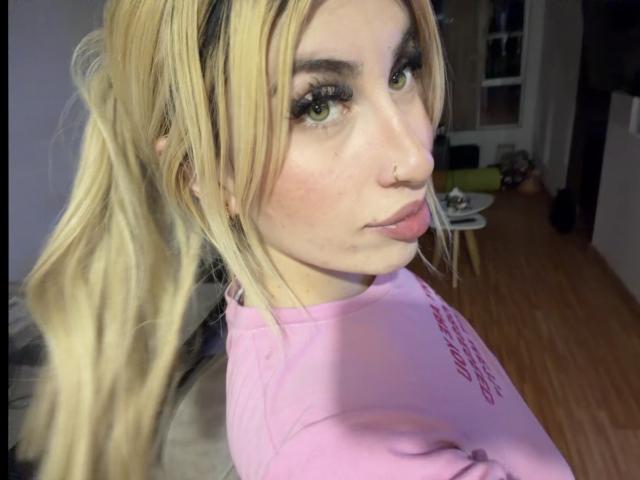 SammyViolet with blonde pigtails, green eyes, and long eyelashes, wearing a pink shirt, looking over her shoulder, nose ring, and slightly pouty lips, in a well-lit room.