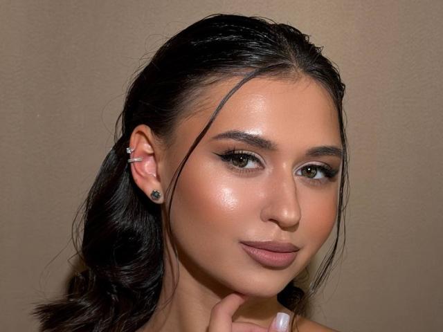 LangCarpinello heeft een glanzende bruine huid, donker, nat haar en draagt een zwarte oorbel. Haar make-up omvat een gevleugelde eyeliner en een mat roze lippenstift. Haar hand raakt haar kin..