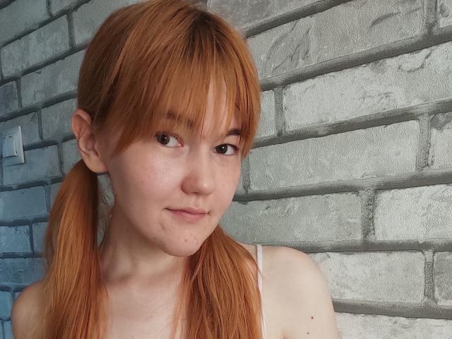 SheritaChieng has red pigtails, a fair skin tone, and small tits, with a subtle smile and freckles, wearing a white top, against a gray brick wall.
